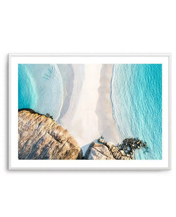 Framed aerial photograph of a coastal beach scene with a white frame and white border. The composition features a white sandy beach curving between turquoise ocean water on the right and a large rocky headland on the left, with gentle waves breaking on the shore.
