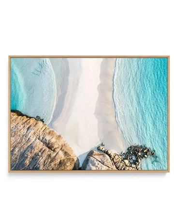 Aerial photography canvas print of a meeting point at Wylie Bay, featuring a pristine white sand beach meeting turquoise ocean water on the right and rugged brown and grey rock formations on the left, with gentle waves creating white foam patterns. The canvas is displayed in a natural wood frame.