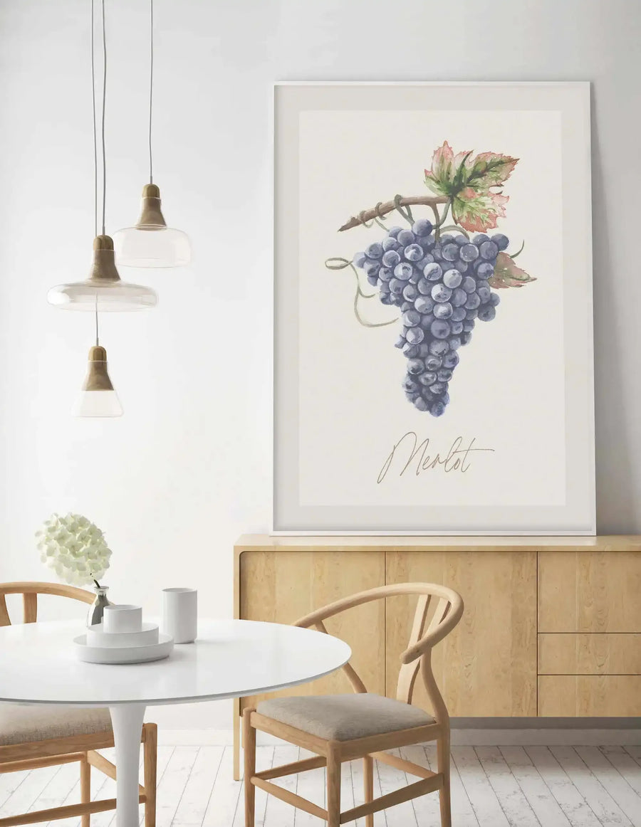 Lifestyle shot of a dining area featuring a framed Merlot art print displayed on a light wood sideboard. The print depicts a cluster of deep purple grapes on a vine with green and reddish-brown leaves, rendered in a watercolor style, with the word "Merlot" in a elegant script below. The room has white walls, a white round table with two light wood chairs, and three glass pendant lights hanging above the table.