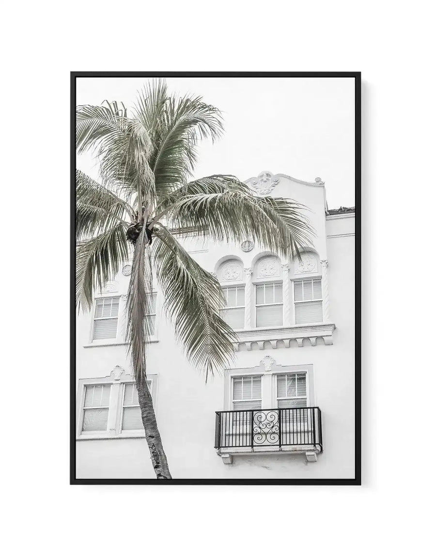 Framed black and white photograph of a palm tree in front of a white building with a balcony, printed on canvas and displayed in a black frame with white border. The composition features a tall palm tree with long fronds on the left, partially obscuring a white multi-story building with ornate architectural details and several windows. A small balcony with a decorative wrought iron railing is visible on the lower right.