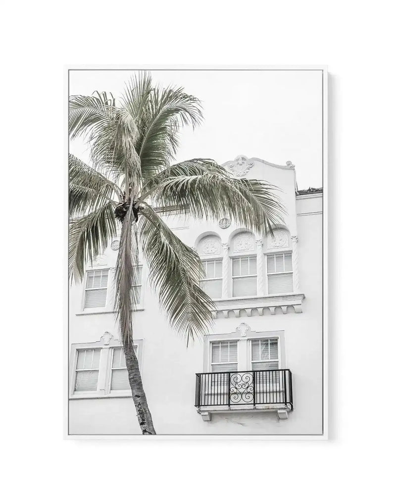 Framed black and white photograph of a white Art Deco building with a tall palm tree in the foreground, with a white frame and white border. The building features arched windows, decorative architectural details, and a small wrought-iron balcony with an ornate scroll design.