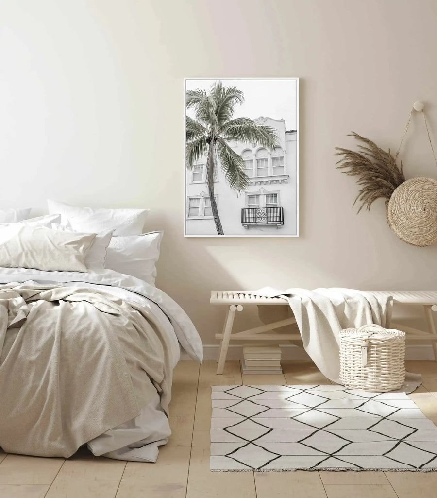 Lifestyle shot of a minimalist bedroom featuring a framed black and white photograph of a palm tree and building. The print is displayed above a light wood bench with a cream throw and a white woven basket. The bedroom has light cream walls, a bed with white and cream bedding, and a white rug with a black diamond pattern on a light wood floor.