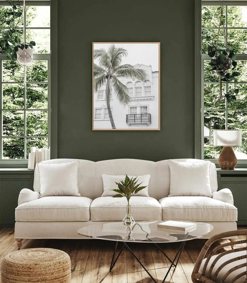 Miami Palms framed canvas art print displayed above a cream linen sofa in a sage green room. The artwork features a minimalist black and white pencil sketch of a tall palm tree in the foreground with a colonial-style white building and balconies in the background. The canvas has a natural wood frame with white border. The print has a soft, monochromatic aesthetic that contrasts with the rich sage green wall, creating a calm, tropical-inspired interior setting.