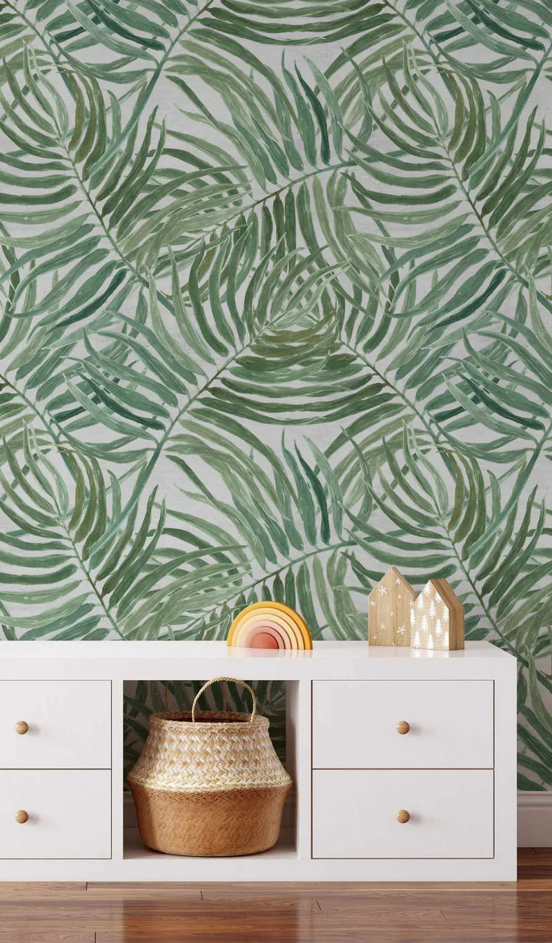 Peel and stick wallpaper featuring a repeating pattern of hand-painted palm fronds in varying shades of emerald green, sage green, and olive green on a soft white background. The botanical design has a loose, organic watercolor quality with visible brushstroke texture. The wallpaper is displayed in a child's room above a white storage unit with wooden knobs, a woven basket, a wooden rainbow toy, and two small wooden house decorations.