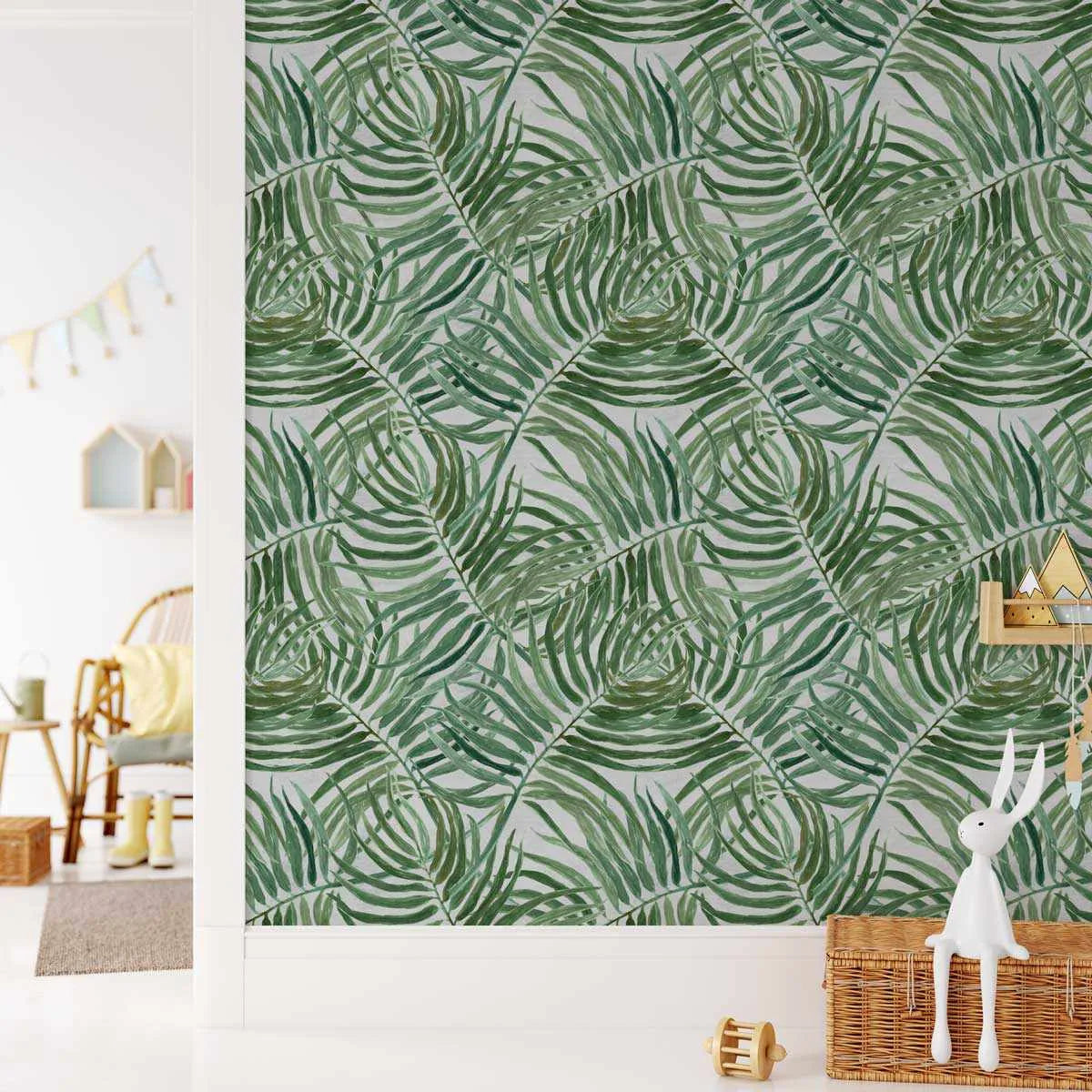 Miami Palms Wallpaper displayed on a bedroom wall featuring a repeating pattern of overlapping green palm fronds in varying shades of forest, sage, and olive green on a light gray background. The botanical illustration style shows detailed line work creating a layered, tropical leaf composition. The wallpaper is shown in a styled room setting with natural wood furniture, wicker storage baskets, a white decorative rabbit figurine, and soft furnishings, creating a calm, nature-inspired interior.