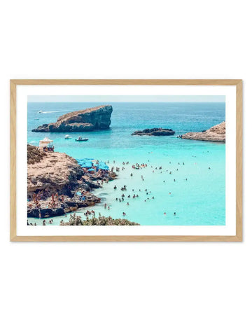 Framed photography art print of a vibrant blue lagoon in Malta, displayed in a natural oak frame with white border. The scene captures a sunny day with numerous people swimming in the turquoise water and relaxing on the rocky shore, dotted with blue umbrellas and a white tent. In the distance, large rock formations rise from the clear blue sea, with a few boats visible on the horizon.