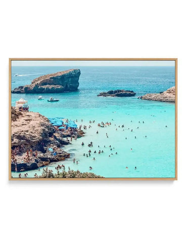 Framed canvas art print depicting a Mediterranean coastal scene in Malta. The photograph shows crystal-clear turquoise water filled with swimmers, rocky tan-colored cliffs and islands, small boats, and a beige beach tent on the left shore. A large natural rock formation dominates the center distance. The image is mounted in a natural wood-toned frame with a horizontal landscape orientation, displaying vibrant blues, sandy browns, and green vegetation along the rocky coastline.