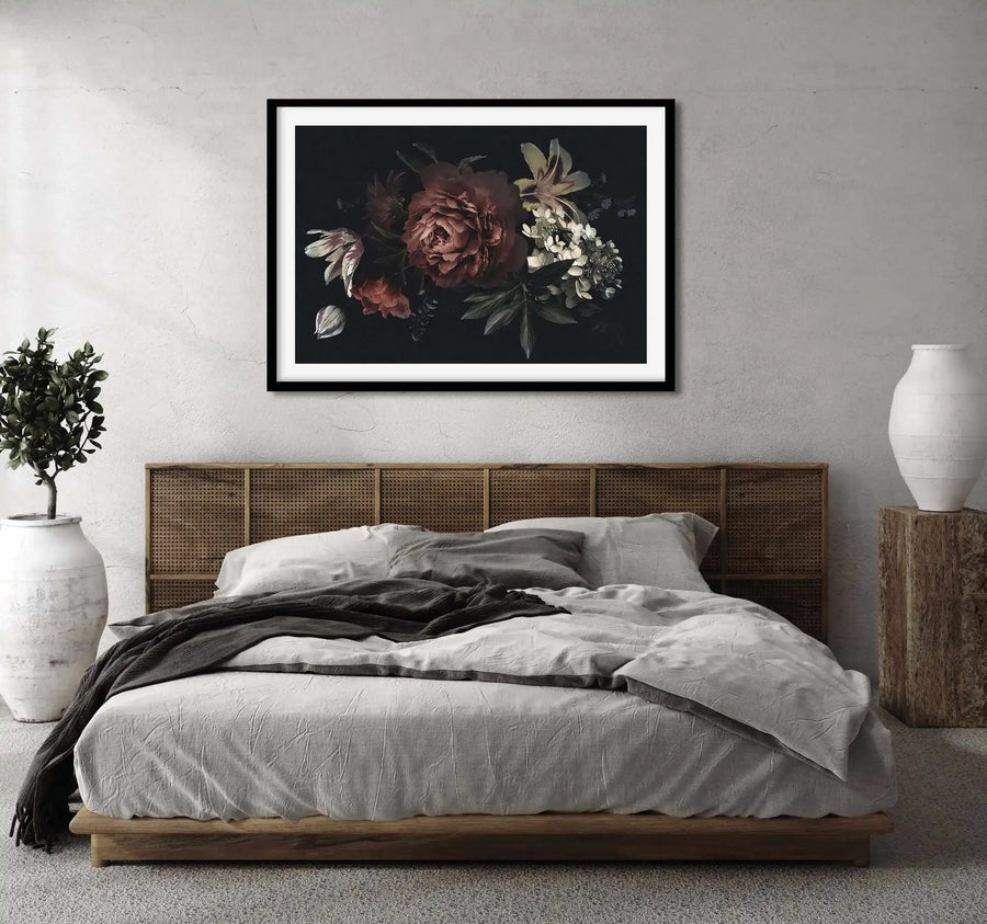 Lifestyle shot of a bedroom featuring a large framed art print above a bed with a woven cane headboard. The print features a dark, moody botanical illustration of deep red peonies, white hydrangeas, and pale green lilies on a black background, with a black frame and white border. The bed has light grey linen bedding and a dark grey throw, with a tall white ceramic vase holding a green plant on the left and another white vase on a wooden block on the right.