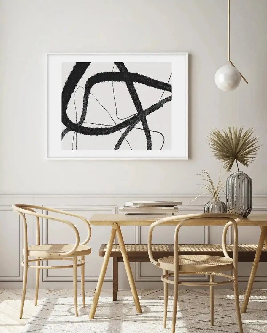 Dining Room:
Large framed abstract art print displayed above a light wood dining table with two chairs in a minimalist dining room. The print features thick, textured black brushstrokes forming an overlapping, dynamic composition on a white background, with a white frame and white border. The room includes a light wood dining table, two light wood chairs with woven seats, a dark wood bench, a brass pendant light with a white globe, and dried palm leaves in a ribbed glass vase.