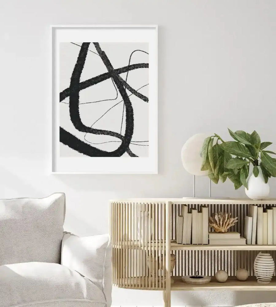 Large framed art print displayed above a curved white sofa in a minimalist living room. The print features bold, abstract black brushstrokes forming an interlocking, fluid design on a white background, with a white frame and white border. The room includes a light wood slatted cabinet with books and decor, and a potted green plant.