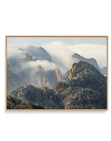 Framed canvas print of a misty mountain landscape, likely Huangshan Mountain in China, with a natural wood frame. The scene features rugged, snow-dusted peaks partially obscured by soft, ethereal clouds and fog, creating a serene and atmospheric composition. The mountains are a mix of dark grey rock and evergreen trees with hints of golden light on some peaks, suggesting sunrise or sunset.