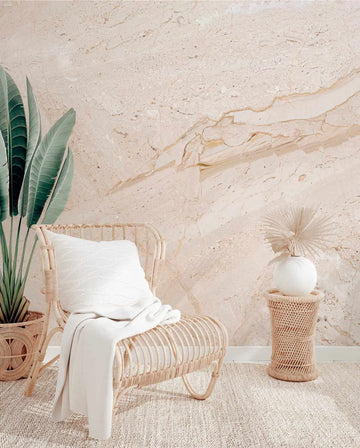 Modern Marble Wallpaper in Blush displayed on a bedroom wall behind a natural woven rattan chair. The wallpaper features an organic marble pattern in soft blush pink, cream, and warm taupe tones with natural veining and fluid swirls throughout. The textured finish mimics polished stone with subtle depth and dimension. The room setting includes natural woven accents, white linens, dried palm leaves, and neutral furnishings that complement the wallpaper's warm, earthy aesthetic.