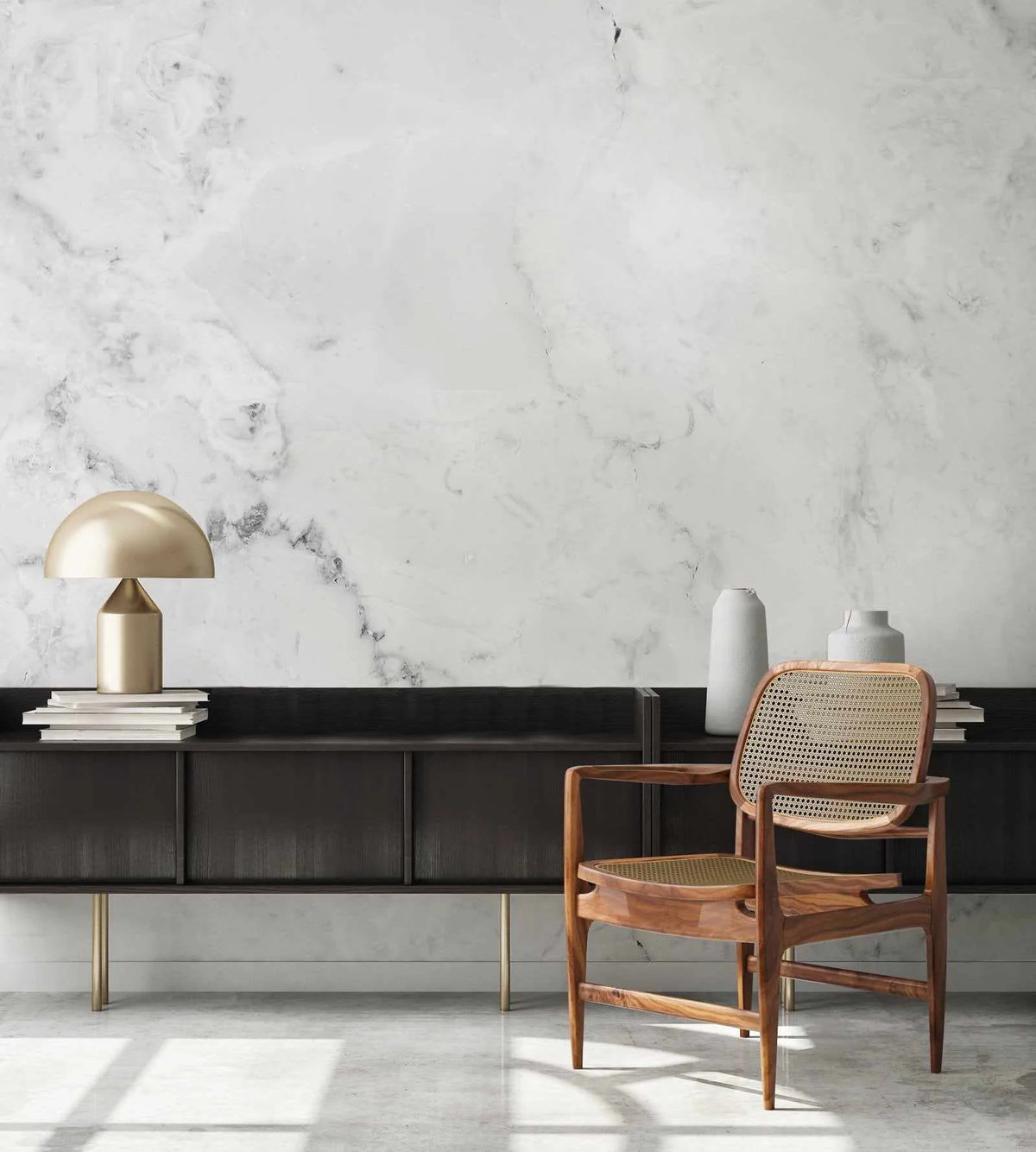 "Modern white and grey marble wallpaper with a subtle, organic vein pattern displayed on a wall above a dark wood sideboard. The room features a gold mushroom-shaped lamp, stacks of books, and two light grey vases on the sideboard, with a wooden chair with cane detailing in the foreground. Sunlight casts shadows on the light grey floor."