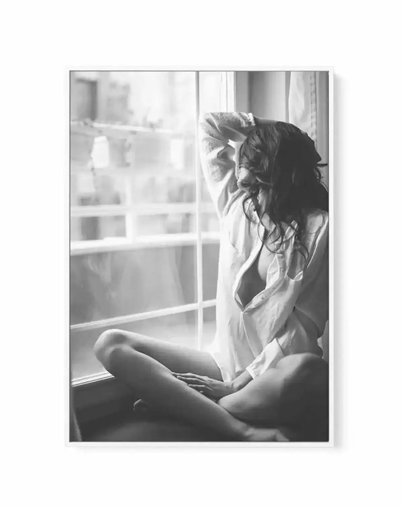 Black and white photograph printed on framed canvas depicting a woman sitting on a windowsill gazing outside. She wears a light linen shirt and has long dark hair pulled back. Bright natural light streams through a multi-paned window behind her, creating soft shadows across her figure. The composition captures an intimate, contemplative mood with fine photographic detail and tonal gradation. Canvas is mounted in a natural wood frame with white border.