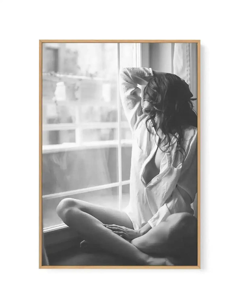 Black and white framed canvas art print titled "Mondays In Paris I" featuring a contemplative woman sitting by a window with grid panes. She wears a light button-up shirt and gazes outward with her hand raised to her head. Natural light streams through the window, creating soft shadows across her face and body. The photograph has a grainy, vintage quality with high contrast between light and dark areas. The canvas is mounted in a natural wood frame with a white border, creating a gallery-style presentation.