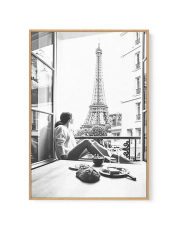 Black and white framed canvas art print titled "Mondays In Paris II" featuring a woman sitting on a window seat looking out at the Eiffel Tower. The photograph shows an open window with metal railings on a balcony, Parisian apartment buildings visible on either side, and a plate with food and bowl on the windowsill in the foreground. The image has a classic, nostalgic quality with soft grey tones throughout. The canvas is mounted in a natural wood frame with a white border.
