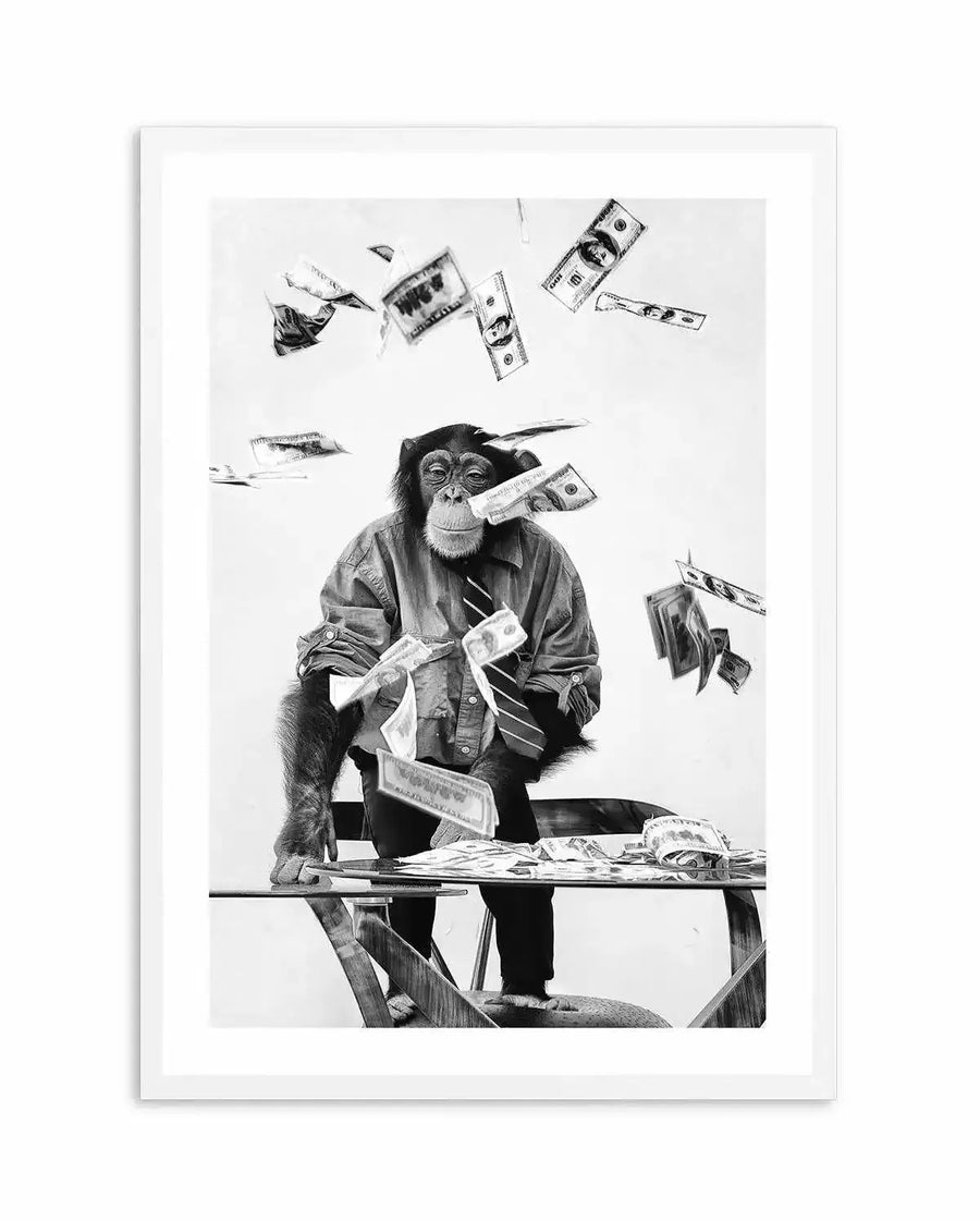 Framed black and white photograph of a chimpanzee in a collared shirt and tie, sitting at a glass table with money falling around it. The chimpanzee holds a bill to its mouth, and more bills are scattered on the table. The background is plain white, with a white frame and white border.