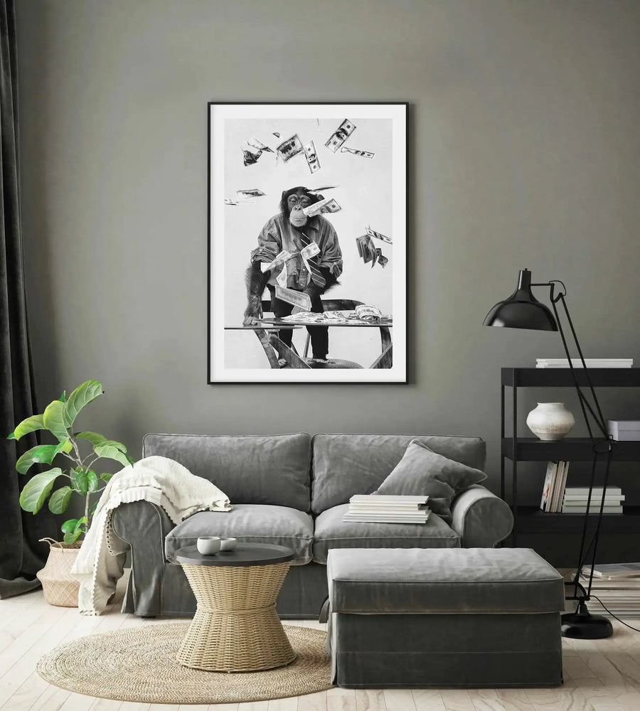 Large framed black and white art print displayed above a grey sofa in a modern living room. The print features a chimpanzee in a shirt and tie sitting at a table with money falling around him. The room includes a grey sofa with a cream throw, a wicker coffee table, a large green plant, and a black floor lamp next to a black shelving unit.
