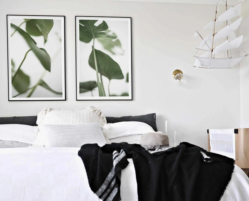 LIFESTYLE SHOT — Bedroom:
Two framed art prints featuring close-up photography of green monstera leaves against a bright white background, displayed side-by-side above a bed in a minimalist bedroom. The prints have black frames and white borders. The bed is dressed in white linen with a black and white striped throw blanket, and a decorative white sailboat hangs on the wall to the right.