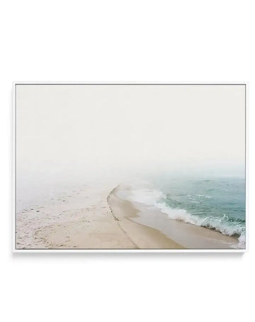 Framed photography canvas print of a misty coastal beach scene with a white frame. The composition features a long stretch of sandy beach curving into the distance, with gentle turquoise waves breaking on the shore under an overcast, hazy sky.