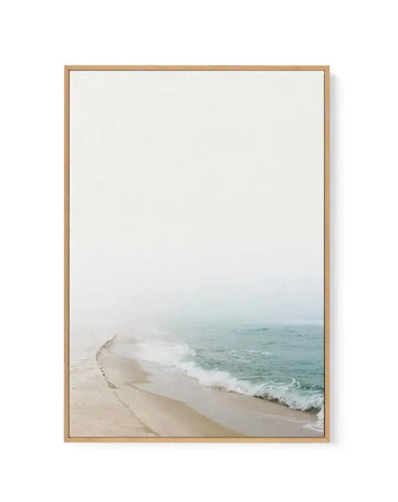 Framed photography print of a misty beach scene with a natural wood frame and white border. The composition features gentle waves breaking on a sandy shore under a pale, overcast sky, creating a serene and ethereal mood.