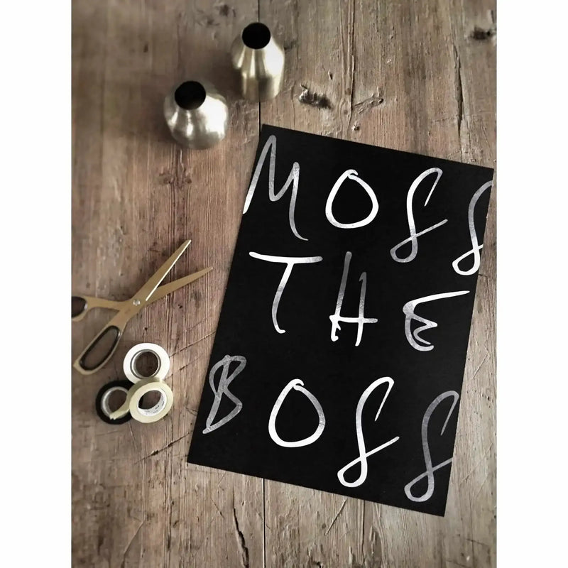 Flat lay styled product shot of "Moss The Boss" art print on weathered gray wood surface. Black paper features handwritten white chalk-style lettering reading "MOSS THE BOSS" arranged in three lines. Surrounding the print are craft supplies including two silver metal bells with black tops, white tape rolls, and gold-handled scissors, creating a styled workspace composition.