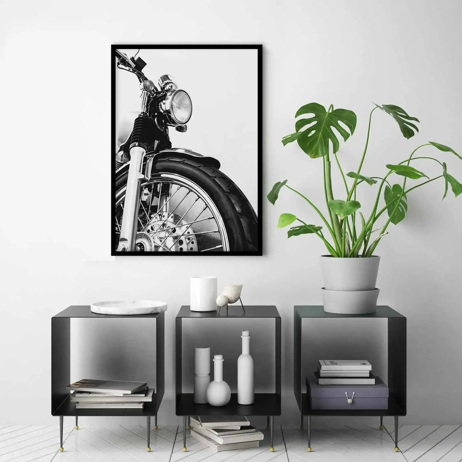 Black and white photograph of a vintage motorcycle displayed in a black frame with white border, showing a close-up angled view of the front wheel, tire, fork, headlight, and handlebars against a light background. The framed print hangs on a white wall above three black open-shelf nightstands with brass legs. The nightstands display white ceramic vessels, books, and a potted Monstera plant with large green leaves in a white pot. Minimalist, monochromatic room styling with a modern aesthetic.