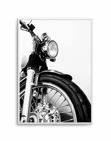 Black and white photograph of a classic motorcycle shown in close-up detail, capturing the front wheel with chrome spokes and black tire, the lower front fork, headlight, and handlebars against a plain white background. The high-contrast monochromatic image emphasizes the metallic shine and mechanical details of the bike. Framed in a light gray frame with white border.