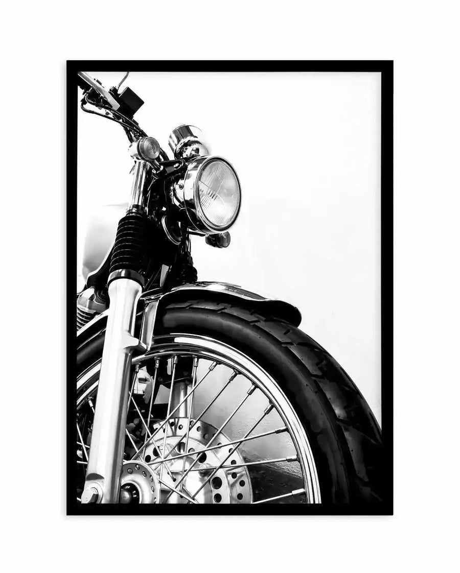 Framed black and white photograph of the front of a vintage motorcycle, displayed in a black frame with a white border. The low-angle composition focuses on the chrome headlight, handlebars, front wheel with spokes, and tire, against a bright white background.