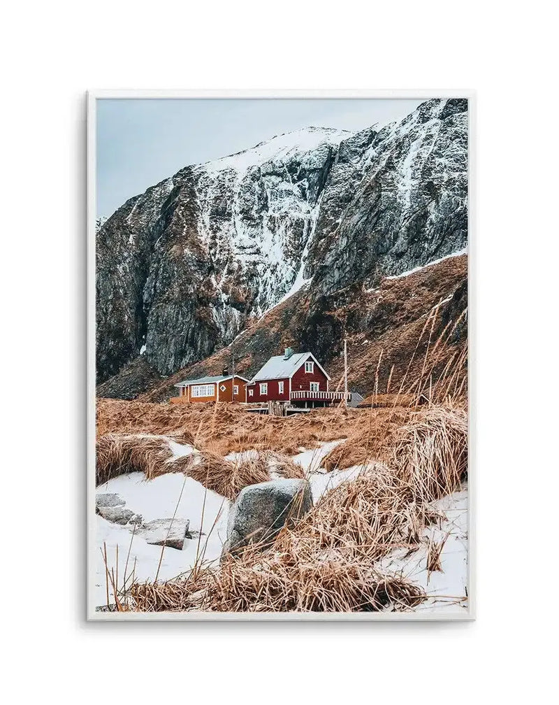 Framed art print featuring a photograph of a red wooden cabin nestled against a dramatic snow-capped mountain cliff. The cabin sits in a barren landscape with brown dried grasses, scattered rocks, and patches of snow in the foreground. Towering dark rocky cliffs with white snow create a striking backdrop. The image has cool tones with contrasts between the warm red building, neutral earth tones, and the cold grey and white mountain. The photograph is mounted in a white frame with white border.