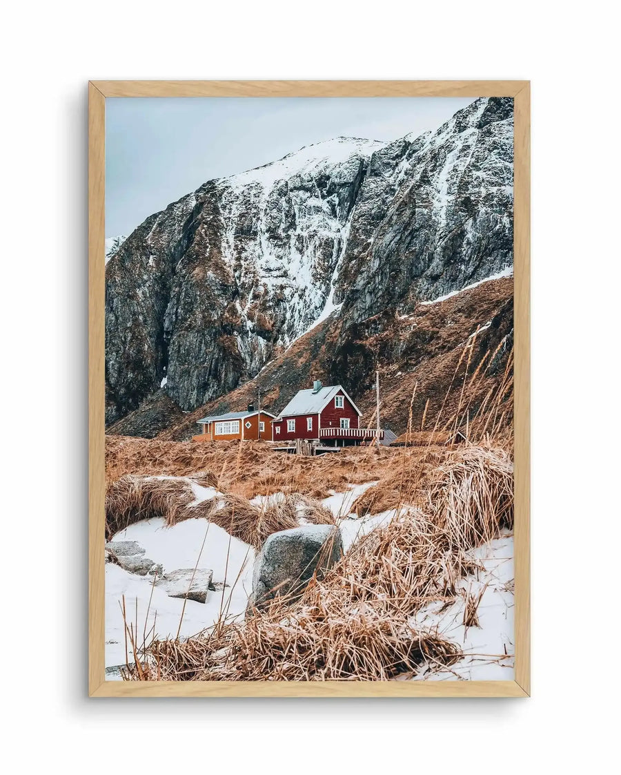 Framed art print featuring a photograph of a red wooden cabin nestled at the base of dramatic snow-capped mountains. The cabin sits in a winter landscape with brown grasses, patches of snow, and scattered rocks in the foreground. Towering dark grey and white rocky cliffs rise steeply behind the structure. The sky is pale blue. The print is displayed in a natural wood frame with white border.
