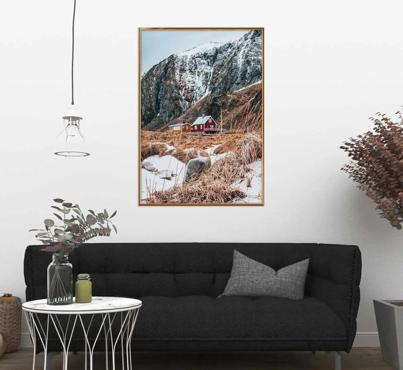 Framed art print displayed above a black sofa in a modern living room. The photograph shows a red wooden cabin nestled at the base of snow-capped mountains, surrounded by brown dried grasses and patches of snow. A small stream runs through the foreground. The image has a natural wood frame with a white border. The room features a round white side table with geometric black patterns, a hanging pendant light, potted plants, and dried botanical decor, creating a minimalist Scandinavian aesthetic.