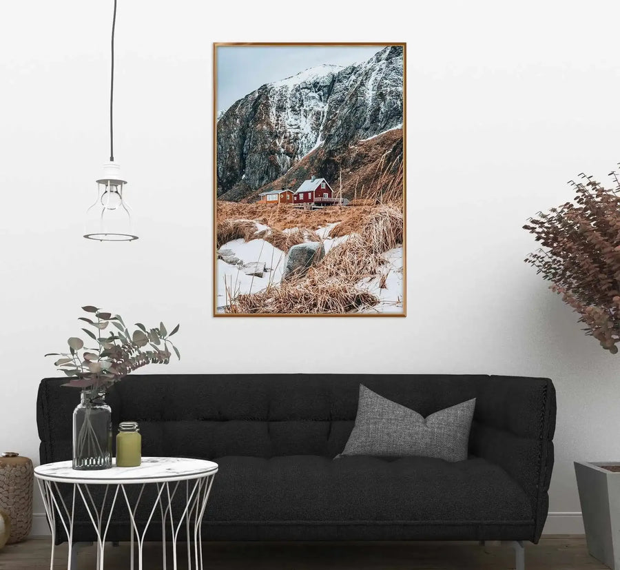 Framed art print displayed above a black sofa in a modern living room. The photograph shows a red wooden cabin nestled at the base of snow-capped mountains, surrounded by brown dried grasses and patches of snow. A small stream runs through the foreground. The image has a natural wood frame with a white border. The room features a round white side table with geometric black patterns, a hanging pendant light, potted plants, and dried botanical decor, creating a minimalist Scandinavian aesthetic.