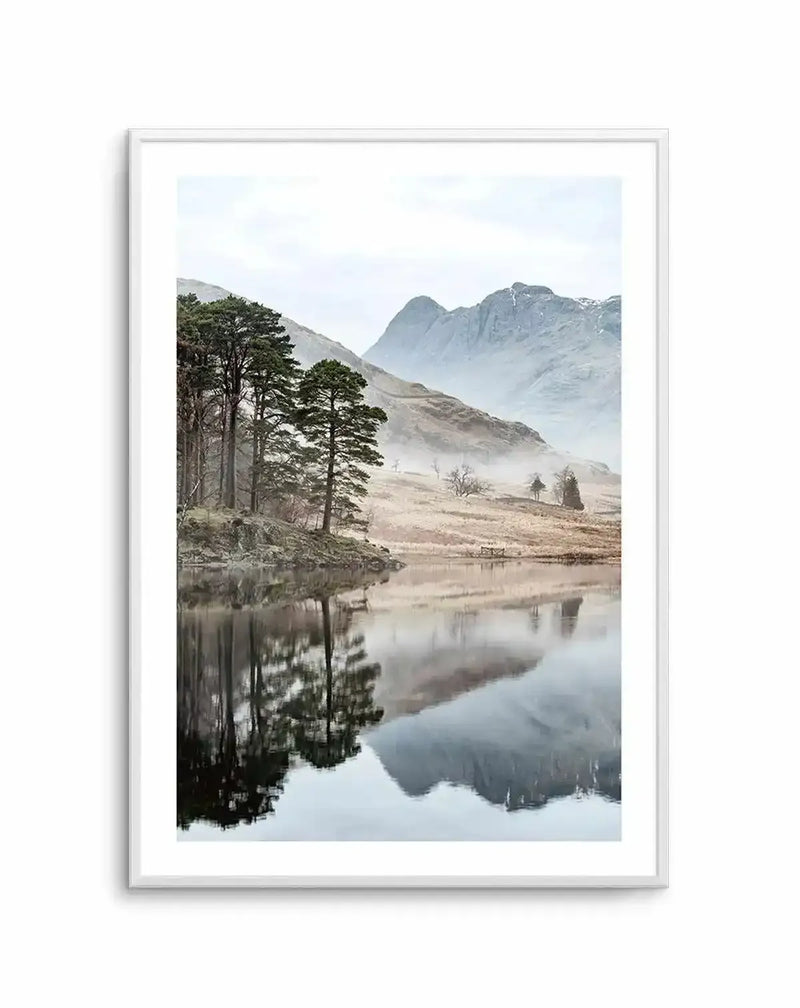 Framed photography art print of a serene mountain lake scene with a white frame and white border. The composition features a still lake reflecting tall pine trees on the left bank and a range of misty, snow-capped mountains in the background under an overcast sky. The foreground shows dry, golden-brown grass and sparse trees on the lake's edge.