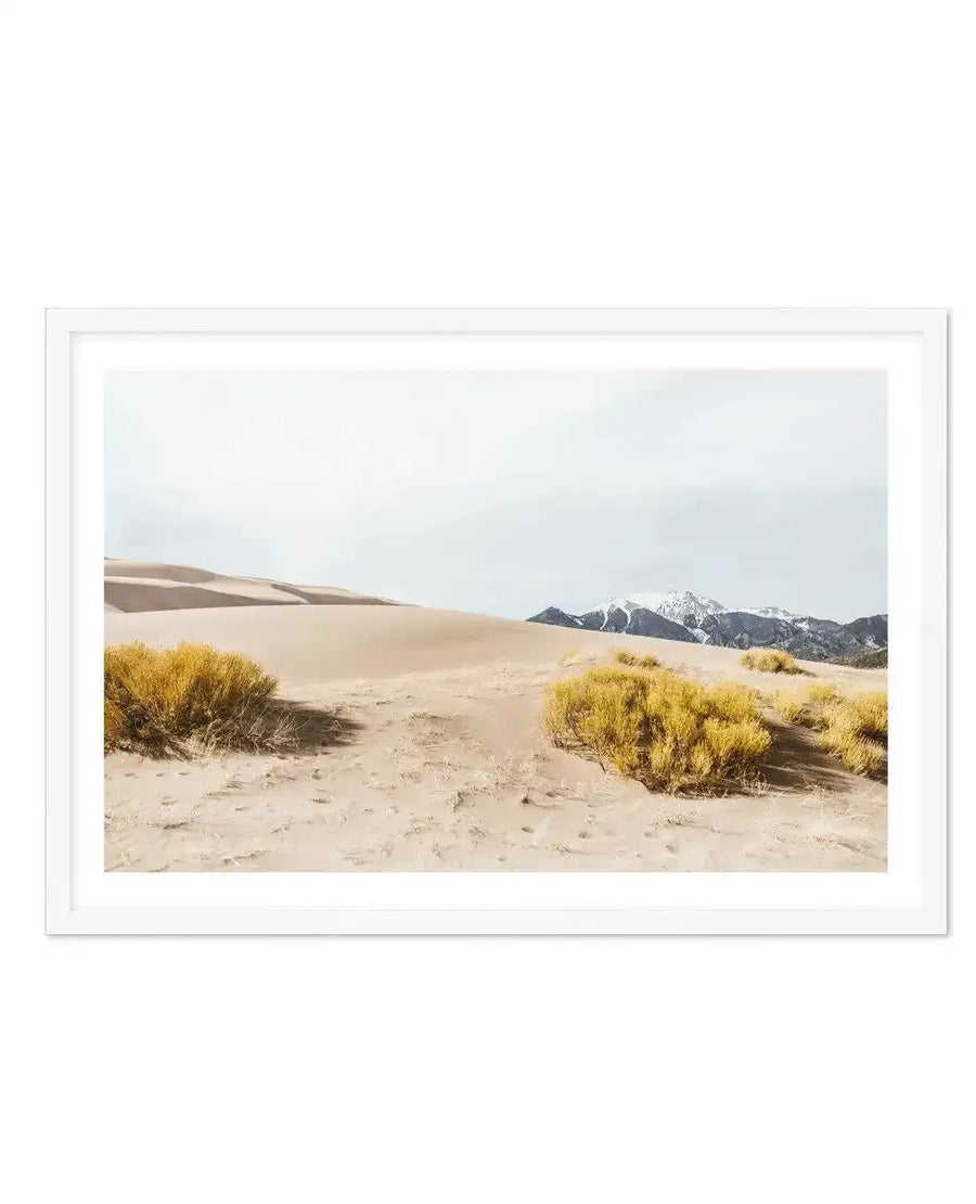 Framed photography art print of a desert landscape with large sand dunes in the foreground and snow-capped mountains in the background, displayed in a white frame with a white border. The sandy terrain is dotted with golden-yellow desert shrubs under a pale, overcast sky.