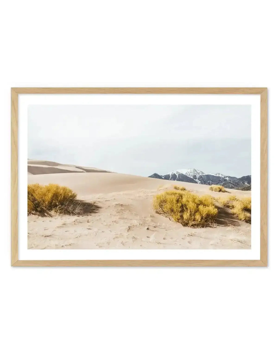 Framed photography art print of a desert landscape with large sand dunes and snow-capped mountains in the distance, displayed in a natural oak frame with white border. The foreground features light brown sand with scattered yellow-green desert shrubs under a pale, overcast sky.
