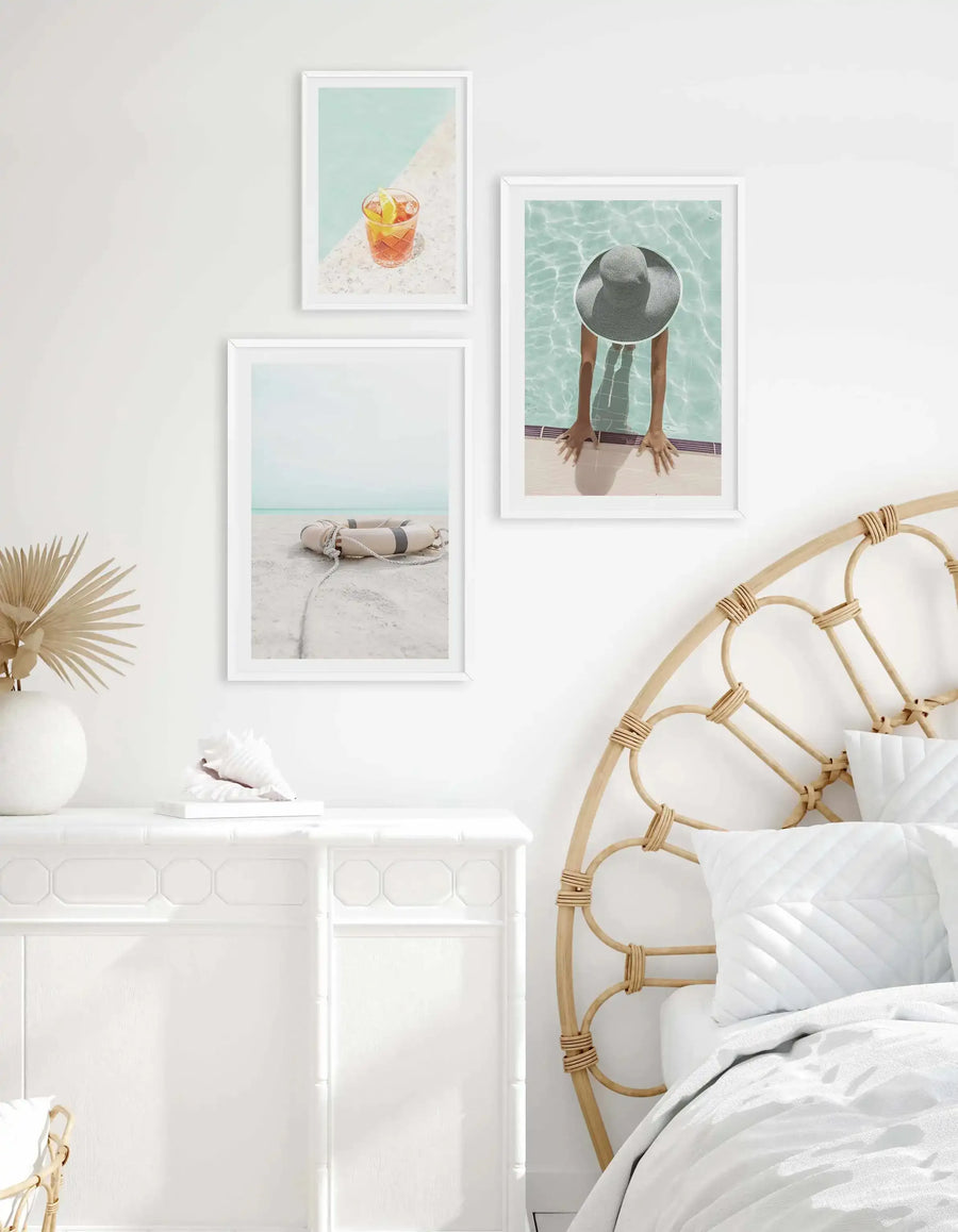 Lifestyle shot of a bedroom featuring three framed art prints above a white dresser. The top print shows a Negroni cocktail on a white tiled surface next to a swimming pool. The middle print shows a person wearing a straw hat with their arms resting on the edge of a swimming pool. The bottom print shows a life preserver on a sandy beach. All prints are framed in white with white borders. The room includes a rattan headboard, white bedding, a white vase with dried palm leaves, and a conch shell.