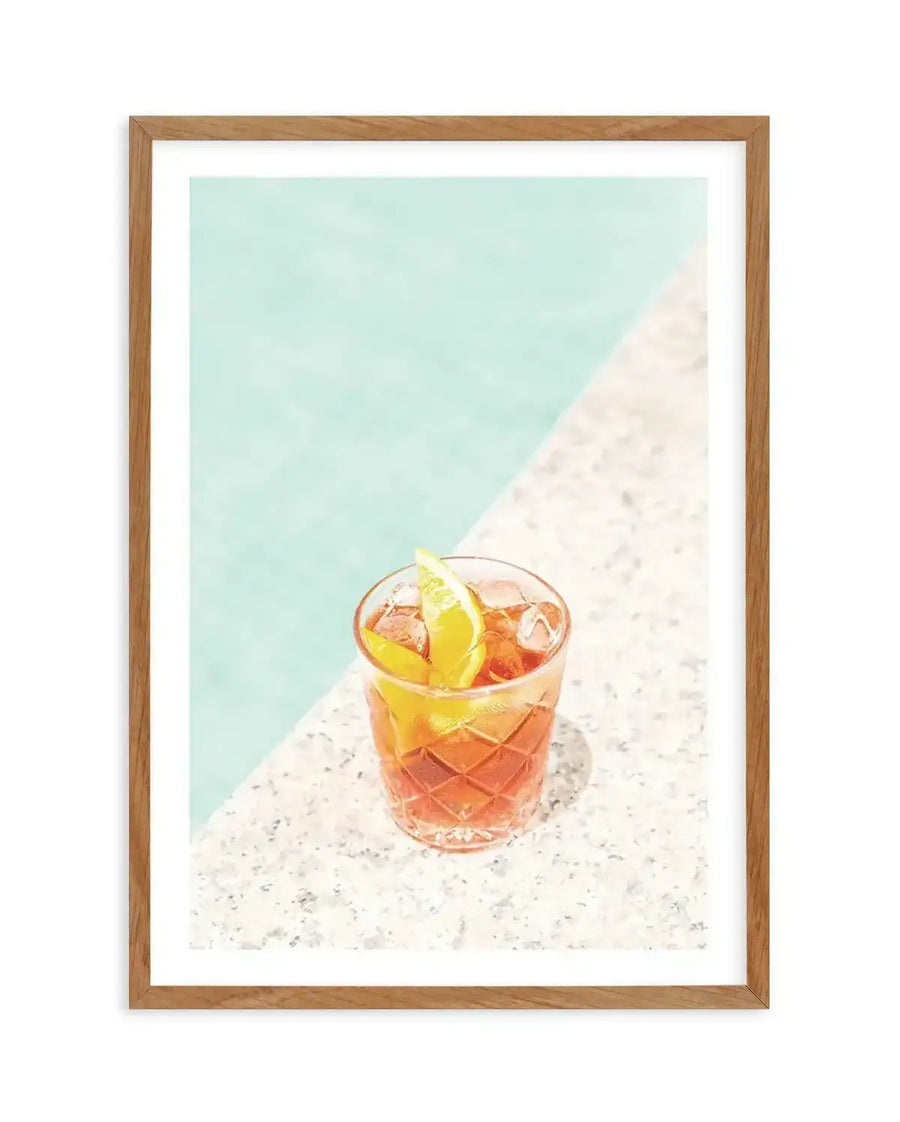 Framed art print featuring a vibrant cocktail in a patterned glass with ice and a lime wedge, set on a speckled white surface next to a turquoise pool. The composition is bright and summery, with a diagonal line separating the pool and the surface. The print is displayed in a natural oak frame with a white border.