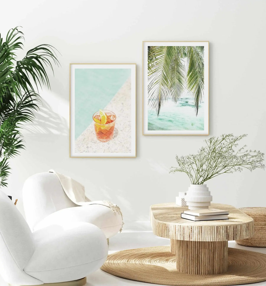 Lifestyle shot of a minimalist living room featuring two framed art prints on a white wall. The left print shows a Negroni cocktail with a lime wedge on a white and grey speckled surface next to a turquoise swimming pool. The right print features a close-up of green palm fronds hanging over clear turquoise water. The room includes a large green plant, a white armchair, a round wooden coffee table with books and a white vase holding white flowers, and a round woven rug.