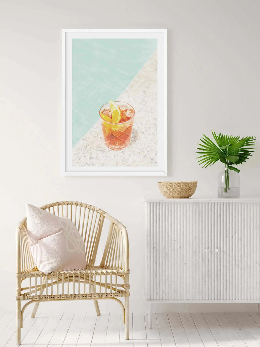 Lifestyle shot of a Negroni art print displayed in a bright, minimalist room. The framed print features a vibrant orange Negroni cocktail with a lime wedge, sitting on a speckled white surface next to a turquoise swimming pool. The print is hung above a white fluted cabinet with a woven bowl and a glass vase with green palm fronds. A light rattan chair with a pale pink cushion sits to the left of the cabinet.
