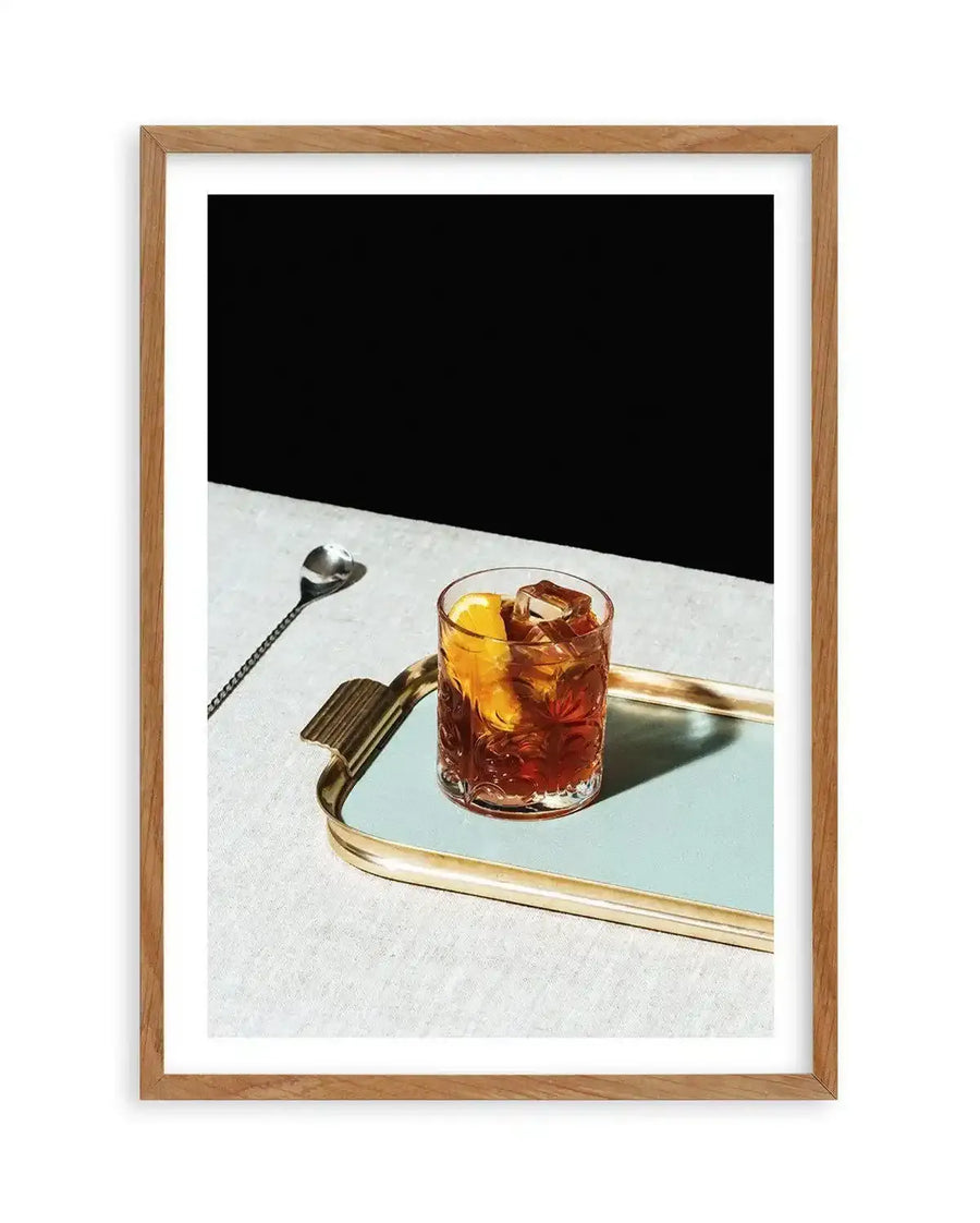 Framed photograph of a Negroni cocktail on a light blue tray, displayed in a natural oak frame with white border. The cocktail, a deep amber liquid with ice cubes and an orange slice, sits in a textured glass. A silver stirring spoon rests on a white linen surface next to the tray, with a stark black background behind.