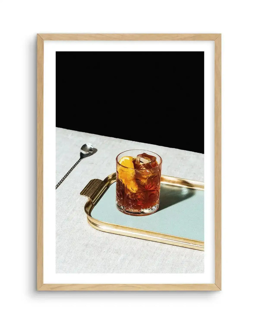 Framed photograph of a Negroni cocktail on a light blue tray with a gold rim, set on a textured white surface against a stark black background, with natural wood frame and white border. The cocktail is in a patterned glass with ice and an orange slice. A silver bar spoon rests to the left of the tray.