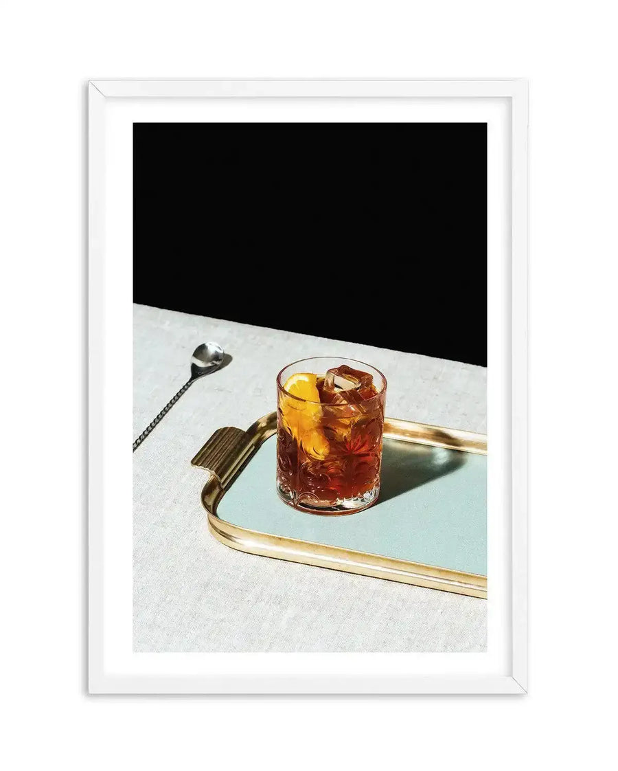 Framed photograph of a Negroni cocktail on a light blue tray with a gold rim, set on a light grey textured surface against a dark black background. The cocktail is in a clear glass with ice and an orange slice, and a long silver stirring spoon rests beside the tray. The print is displayed in a white frame with a white border.