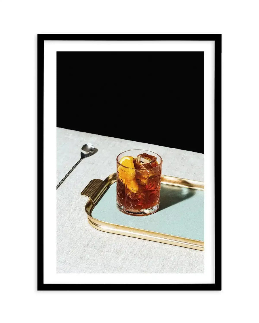 Framed photography art print featuring a Negroni cocktail on a light blue tray, with a black frame and white border. The cocktail, a rich amber color with ice cubes and an orange slice, sits on a reflective light blue tray with gold edges and handles. A silver bar spoon rests on a textured light grey surface to the left, with a stark black background behind the cocktail and tray.