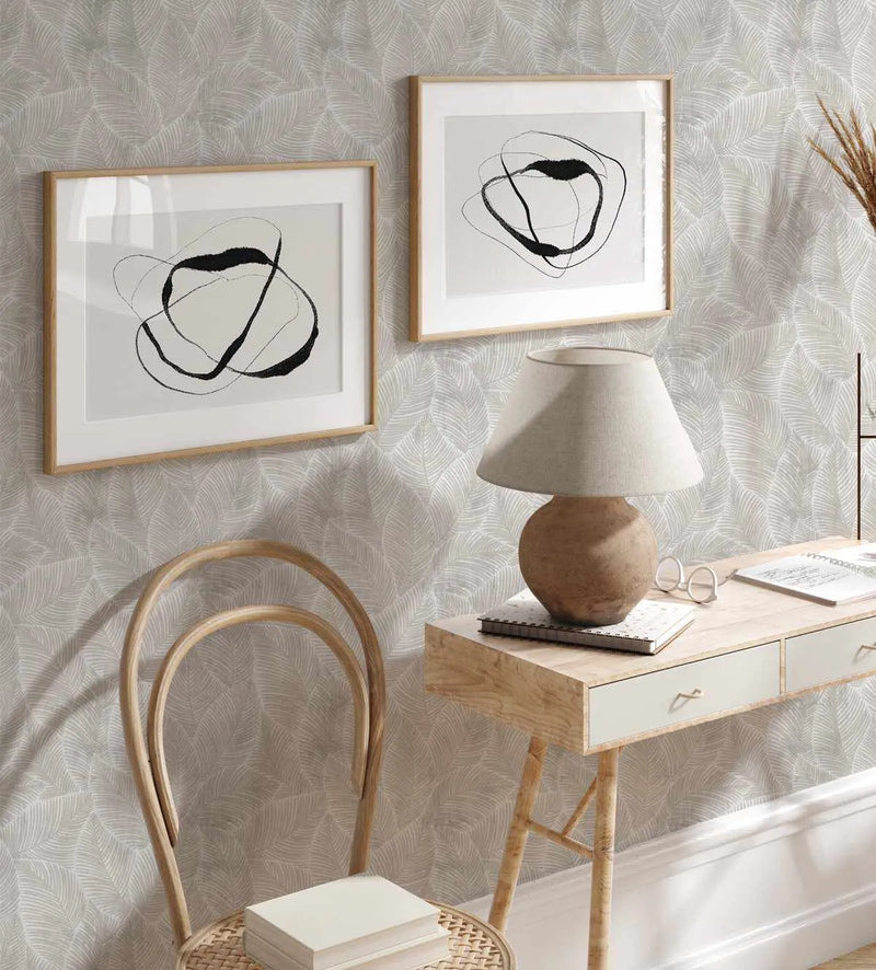 Neutral Leaves Wallpaper displayed on a bedroom wall behind a wooden desk. The wallpaper features a repeating pattern of delicate, feathery palm fronds in soft greige and cream tones with a subtle woven texture. Two framed abstract line drawings with wooden frames hang above the desk. A wooden bentwood chair with woven rattan seat sits in the foreground, and a spherical ceramic table lamp with cream linen shade sits on the desk beside notebooks and eyeglasses, creating a calm, minimalist workspace.