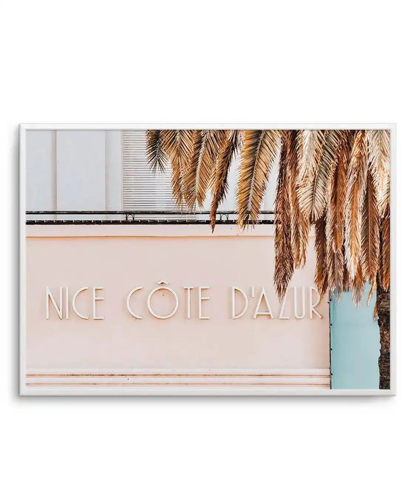 Framed photography art print featuring a light pink building with the words "NICE CÔTE D'AZUR" in white 3D letters, partially obscured by the fronds of a large palm tree in the foreground. The building has a balcony with a black railing, and a light blue wall is visible to the right. The print is displayed in a white frame with a white border.
