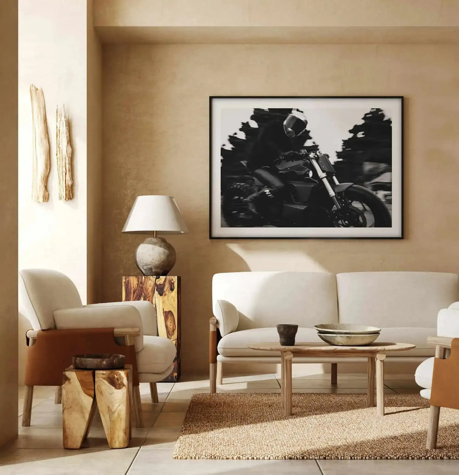 Black and white photograph of a motorcycle rider in motion, mounted in a black frame with white border, displayed on a warm beige wall above a cream linen sofa in a minimalist living room with natural wood accents and neutral furnishings.