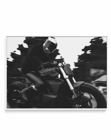 Framed canvas art print featuring a black and white photograph of a person riding a motorcycle at speed, creating a blurred background of trees and a clear focus on the rider and bike. The rider wears a full-face helmet and dark clothing, leaning forward over the handlebars. The motorcycle is dark-coloured with visible front forks and a front wheel in motion. The print is displayed in a white frame.