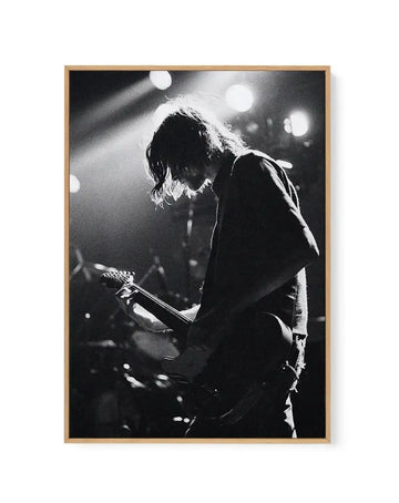 Framed black and white canvas print of a musician playing guitar on stage under bright spotlights. The musician, with long, dark hair, is silhouetted against the light, facing right and looking down at their guitar. The image captures a dynamic moment with visible light rays and a grainy texture, displayed in a natural oak frame.