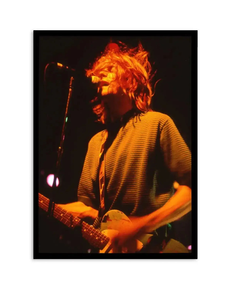 Framed photography art print featuring a dynamic, close-up shot of a musician with long, bright orange hair mid-performance, illuminated by warm stage lights. The musician, seen from the chest up, is wearing a horizontally striped shirt and playing an electric guitar, with their head tilted back and hair flying. The background is dark with soft, out-of-focus lights. The print is displayed in a black frame.